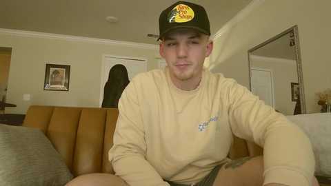 A video of a young Caucasian man with light skin, wearing a beige sweatshirt and green baseball cap, sitting on a bed in a beige room with framed art on the walls.