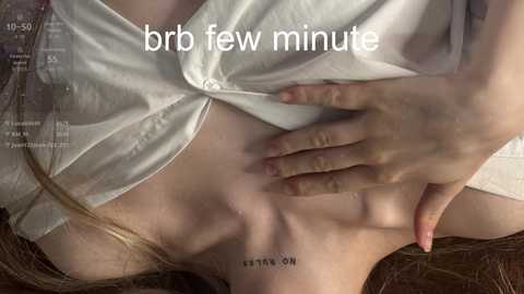 A close-up video of a fair-skinned woman lying on a bed, wearing a white, wet, button-up shirt, with her hand on her stomach, revealing a tattoo that says \"ON.\