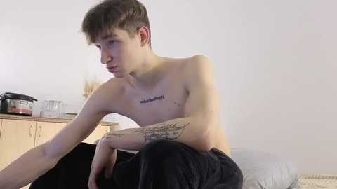 Video of a shirtless young man with short, dark hair, tattoos on his arms and chest, sitting on a bed in a minimalist bedroom with light-colored walls and kitchen items on a wooden cabinet in the background.