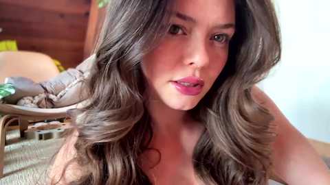 Media: Video of a young woman with long, wavy brown hair and fair skin, wearing pink lipstick, smiling slightly. She's indoors, seated, with wooden furniture and a plant in the background.