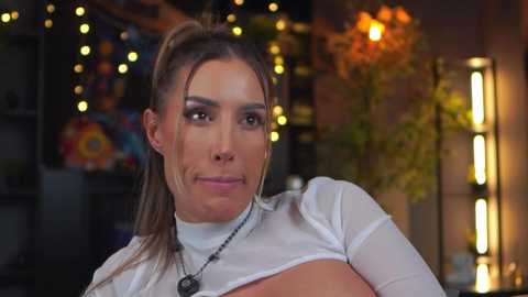 Video of a fair-skinned woman with brown hair in a ponytail, wearing a white crop top, smiling, in a dimly lit room with colorful wall art and a lamp.