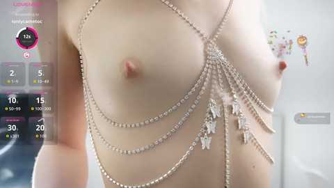 A close-up video of a topless, light-skinned woman with small breasts adorned with a delicate, silver chain choker featuring crystal pendants. The background shows a kitchen with a white fridge and a calendar overlay.