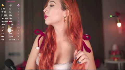 Video of a fair-skinned woman with long, wavy, fiery red hair, wearing a lacy white bra, in a dimly lit room with a pink chair and a blurry background.