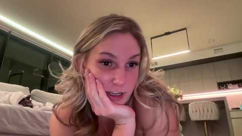 Media: Video of a blonde woman with wavy hair, resting her chin on her hand, in a modern, minimalist bedroom with white furnishings, ambient lighting, and a sleek design.