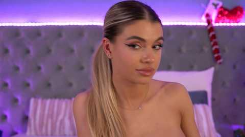 Video of a topless woman with fair skin, long blonde hair in a high ponytail, wearing bold makeup and a delicate necklace, seated on a bed with a tufted gray headboard and festive red and white striped pillow.