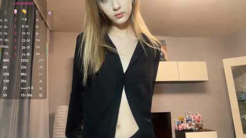 Video of a slender, fair-skinned, young woman with long blonde hair, wearing an unbuttoned black shirt, standing in a minimalist bedroom with beige walls, a white shelf, and a hanging calendar.