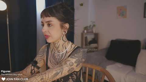 Media: Video of a tattooed, dark-haired woman with piercings, wearing a sheer black top, sitting in a modern living room with a white couch and artwork.