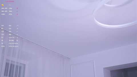 A video of a minimalist, white room with a circular chandelier, white walls, and sheer curtains partially covering a window. A digital display shows system information and temperature, indicating a modern, clean interior.