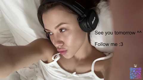 Video of a young woman with light skin and brown hair, wearing a white camisole and black headphones, lying on white bedding, making a kissing gesture. Text on the right reads, \"See you tomorrow, Follow me <3.\
