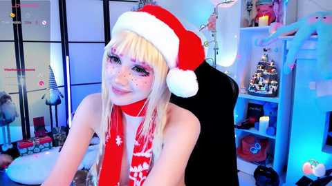 Video of a fair-skinned, blonde woman with freckles, wearing a red Santa hat, white snowflake scarf, and minimal makeup, in a festive, brightly lit room with Christmas decor.