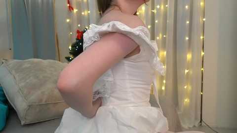 Video of a fair-skinned woman with brown hair, wearing a white, off-the-shoulder, frilled dress, standing in a room with white curtains and fairy lights, holding a blue object.
