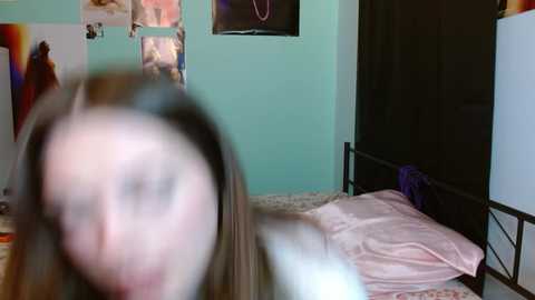 Video of a blurry woman with long hair, standing in a pastel-colored bedroom with a bed, wall art, and a black door.