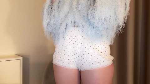Video of a person's lower back and buttocks in white polka-dotted shorts, wearing a white, feathered top. Background features beige walls and a white dresser.