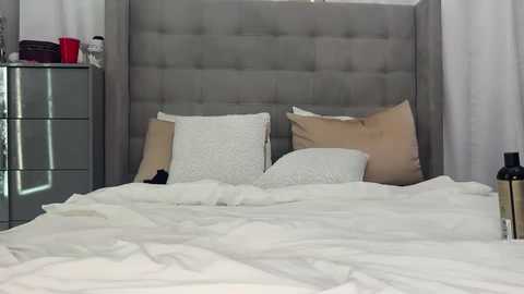 A video of a modern bedroom featuring a gray upholstered headboard, white bedding, beige pillows, a glass nightstand with red and black items, and white curtains.