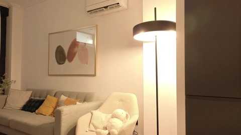 A cozy, modern living room with a beige sofa adorned with pillows, a tall floor lamp, and a white armchair. The room is warmly lit, with a white air conditioner and abstract art on the wall.