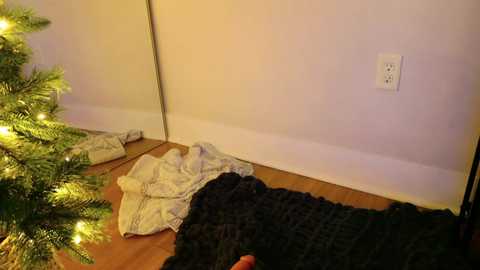 Video of a cozy, lit Christmas tree with a knitted blanket, a folded white blanket, and a white electrical outlet on a wooden floor, all in a dimly lit room.