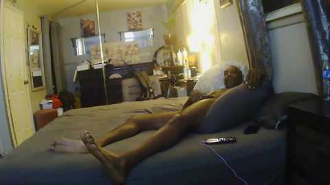 Video of a dark-skinned man lying nude on a bed, surrounded by cluttered, dimly lit room with mirrors, TV, and scattered items.