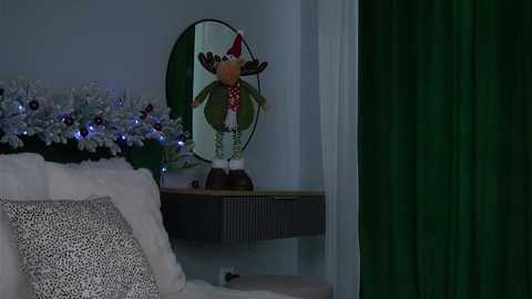 A video of a cozy living room decorated for Christmas with a plush white sofa, a festive wreath, and a green curtain. A wooden shelf holds a wooden Santa figurine, illuminated by a circular mirror.