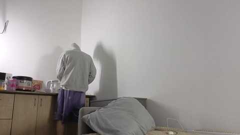 Video of a man in a gray hoodie and purple shorts, standing at a kitchen counter, facing away, casting a shadow on a white wall. Background includes kitchen appliances and a couch with a blanket.