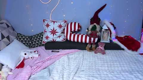 Video of a cozy, festive bedroom with a white bedspread, red and white striped pillows, a stuffed elf, Christmas lights, and a Santa hat, creating a whimsical holiday atmosphere.