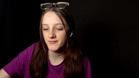 Video of a young girl with long brown hair, wearing a purple T-shirt and white-framed glasses, resting against a black background. She appears contemplative with a slight smile.