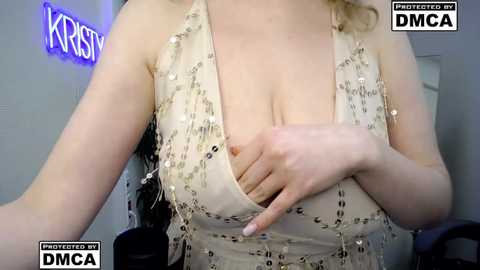 Video of a fair-skinned woman with medium-length blonde hair, wearing a revealing, sequined beige halter top, covering her cleavage with her hand. Background features a neon \"Krispy\" sign and a DMC logo.