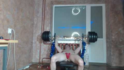 Video of a muscular man in red underwear lifting a barbell with blue weights, seated on a bench in a bare, unfinished room with exposed walls and windows.