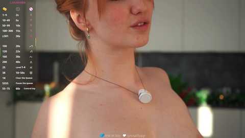 A video of a pale-skinned, freckled woman with red hair in a bun, wearing a white necklace, in a modern kitchen with blurred appliances.