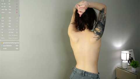 Video of a topless woman with long dark hair, slender build, and multiple tattoos on her left arm, stretching. She wears blue jeans. Background: calendar, lamp, green plant.