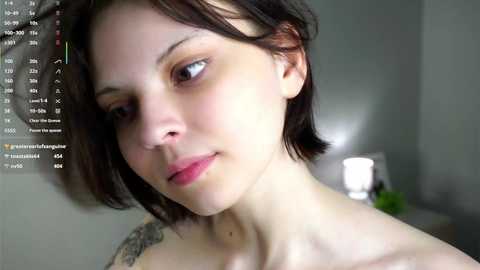 Video of a fair-skinned woman with short brown hair, light makeup, and a subtle smile. She has a small tattoo on her left shoulder. The background is a blurred indoor setting with a lamp.