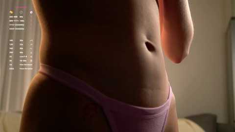 Media: A close-up video of a woman's lower torso in a light pink thong, with a small tattoo on her hip. The background shows a dimly lit room with soft lighting.