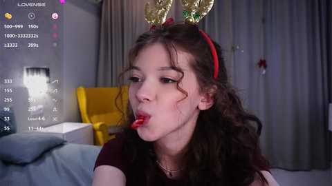 Media: Video of a young woman with curly brown hair, wearing red lipstick, and gold antlers, licking a lollipop in a dimly lit room.