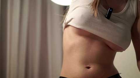 A close-up video of a fair-skinned woman lifting her beige ribbed crop top, revealing her small to medium-sized breasts. She has a flat stomach and wears a black dog tag necklace. Background shows beige curtains and a white ceiling lamp.