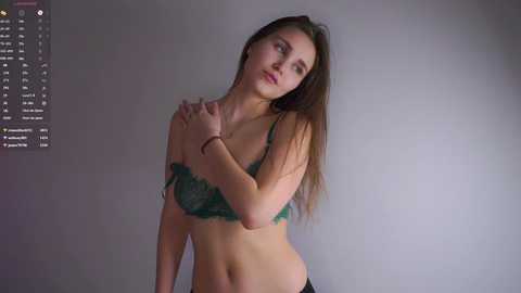 Media: Video of a young Caucasian woman with long brown hair, wearing a green lace bra, standing against a plain gray background. She has a slender physique and a neutral expression.