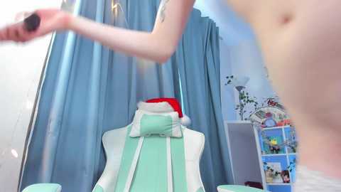Video of a light-skinned person with a tattoo, topless, holding a bottle, in a room with a white mannequin draped in green, blue curtains, and a festive, colorful backdrop.