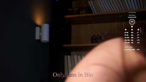 Video of a dimly lit room with a wooden bookshelf, a wall-mounted lamp, and a person's hand holding a smartphone displaying a bio app. Text overlay reads \"OnlyFans in Bio.\