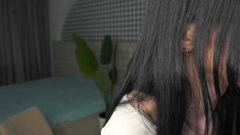 A close-up video of a person with long, black hair, partially covering their face, wearing a white top. The background features a checkered wall, a green plant, and a light blue cushioned bench.