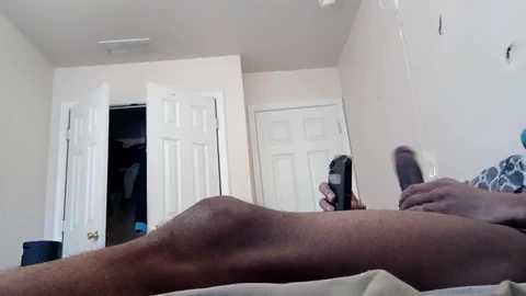 Media: Video of a man lying naked on a bed, holding a black remote, with white doors and beige walls in the background.