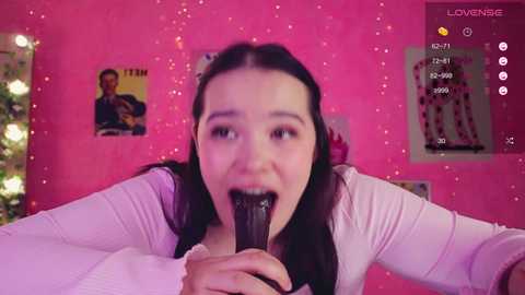 A video of a young woman with long black hair, wearing a white top, licking a black dildo in a pink-themed room with posters and a Christmas tree.