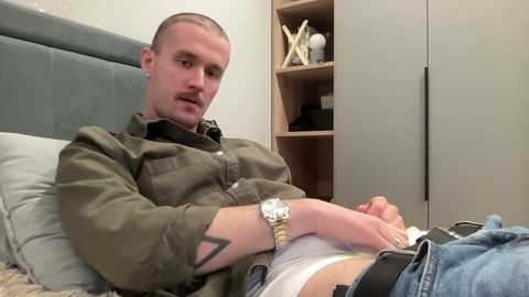 Video of a bald man in a gray hoodie and jeans, lying on a bed with white sheets, holding his crotch, wearing a watch, in a sparsely furnished room with a wooden shelf and white walls.