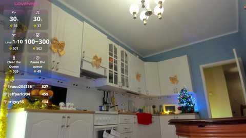 Video of a cozy, modern kitchen with white cabinets, gold ribbon decorations, and a lit Christmas tree in the corner.