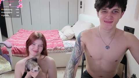 Video of two young women in a bedroom: one topless with fair skin and red hair, the other shirtless with a tattooed arm, both smiling.