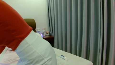 Video of a person wearing a red shirt and light blue pants, lying on a bed with white sheets and a beige pillow, in a room with light blue curtains and a nightstand with a lamp and items.