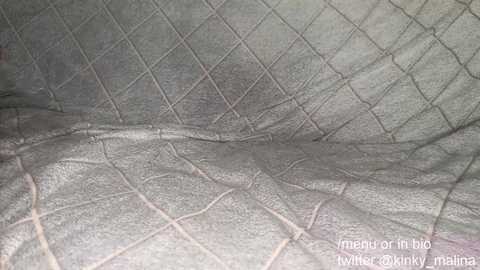 Video of a textured, quilted gray fabric with a diamond pattern, displaying subtle wrinkles and creases. The background is plain, and the image includes a watermark reading \"mayis on bio twitter @coxxminky_malina.\