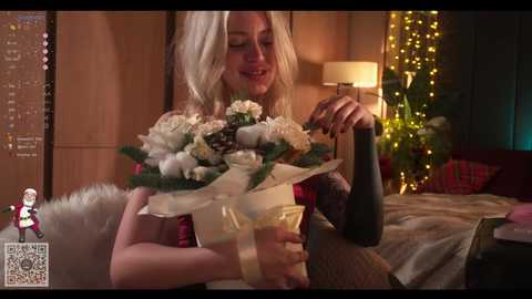 Video of a smiling blonde woman holding a bouquet and a champagne bottle in a cozy, warmly lit bedroom with fairy lights and a bed.