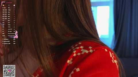 Video of a woman with long, straight, light brown hair, wearing a red sweater with white geometric patterns, seen from the side. Background shows a blue window and dark curtains.