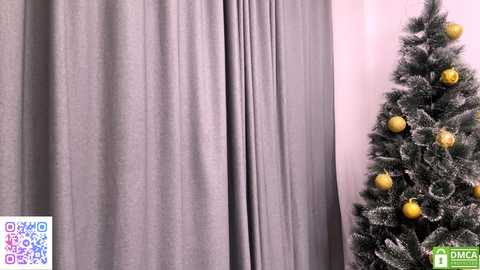 Video of a minimalist room with a gray curtain and a decorated Christmas tree adorned with golden ornaments, set against a pale pink wall.