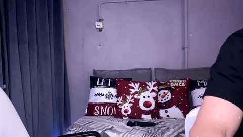 Video of a modern bedroom with a gray wall, a person wearing a black shirt, and a bed with red and white holiday-themed pillows featuring reindeer and snowflakes.