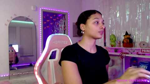 Video of a young woman with light skin, dark hair, and a black top seated in a pink and white gaming chair in a pastel-themed room with neon lights, toys, and a colorful wall mural.