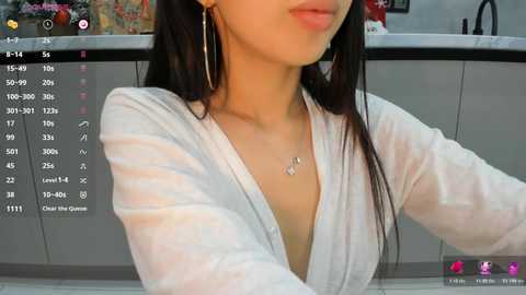 Video of a young Asian woman with long black hair, wearing a white V-neck top and silver hoop earrings, seated in a modern kitchen, with a camera app interface overlay.